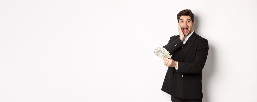 Image of rich and happy man in black suit, winning prize, holding money and looking excited at camera, standing over white background.