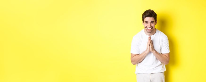 Handsome guy saying thank you, bowing and holding hands in namaste gesture, express gratitude, standing over yellow background.