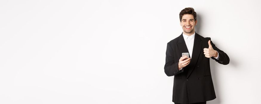 Handsome guy in trendy suit making compliment, showing thumbs-up while using mobile phone, recommending an app or online shop, standing over white background.