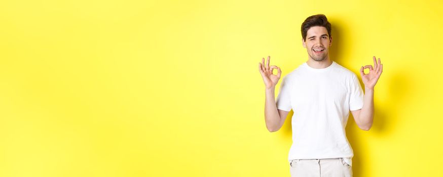 Confident handsome man winking, showing okay signs in approval, like something good, standing over yellow background.