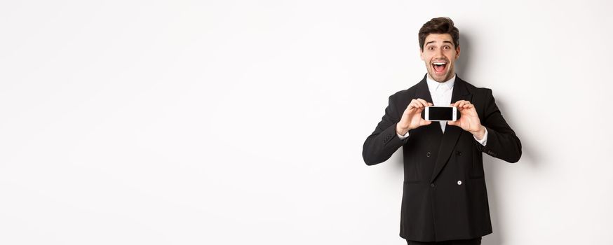 Image of cheerful, handsome man in black suit, showing smarthone screen and looking amazed, standing against white background.