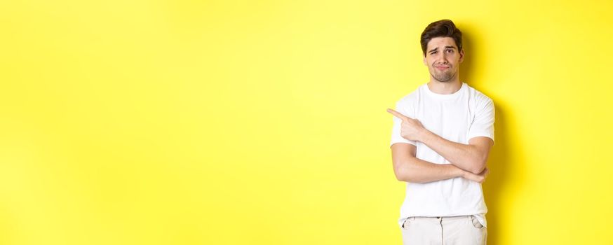 Skeptical and reluctant guy in white t-shirt, pointing finger left and smirking unamused, showing bad advertisement, standing over yellow background.