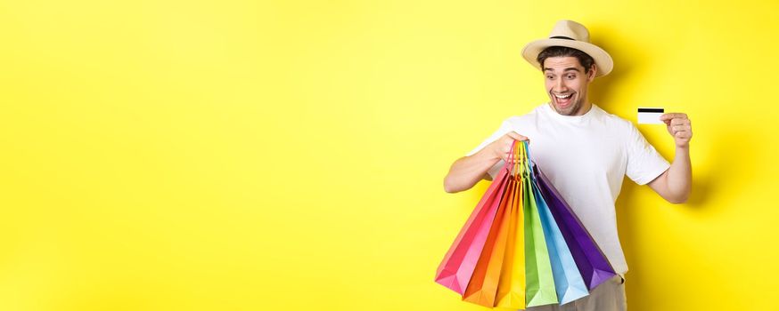 Concept of vacation and finance. Happy man shopper looking at shopping bags satisfied, showing credit card, standing against yellow background.