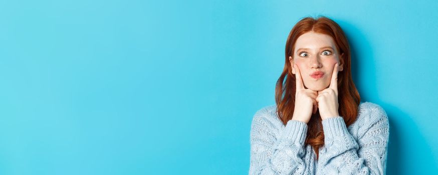 Close-up of funny redhead teen girl making faces, squinting and pocking cheeks, standing against blue background.