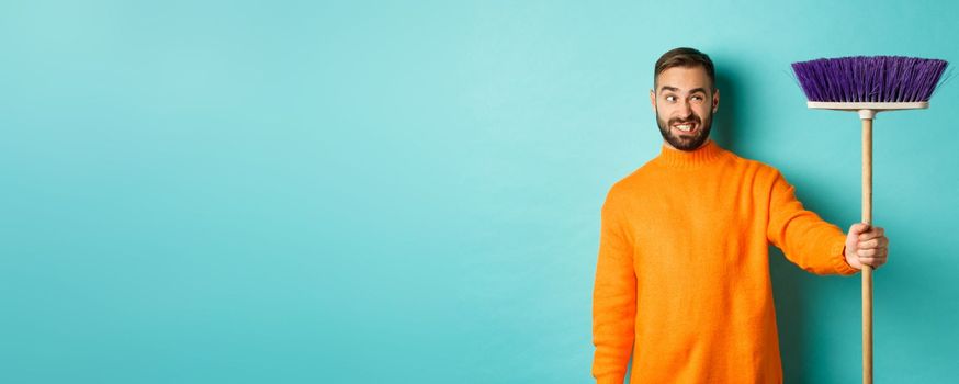 Boyfriend looking confused at broom, forced clean house, doing chores, standing displeased against light blue background.