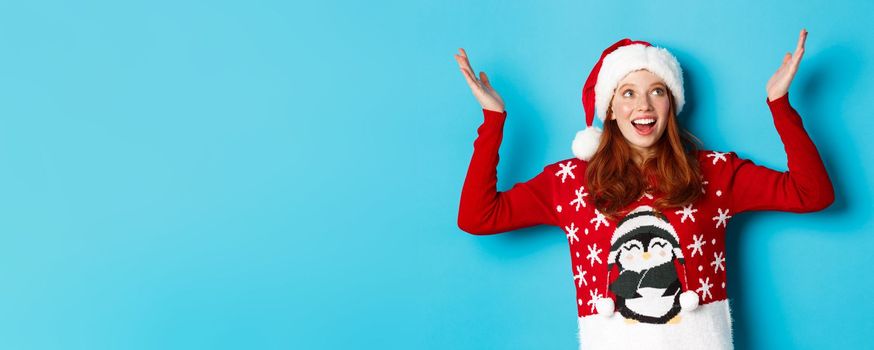 Happy holidays and Christmas concept. Excited redhead girl rejoicing of something falling on her, raising hands up delighted and smiling, wearing Santa hat with xmas sweater, blue background.