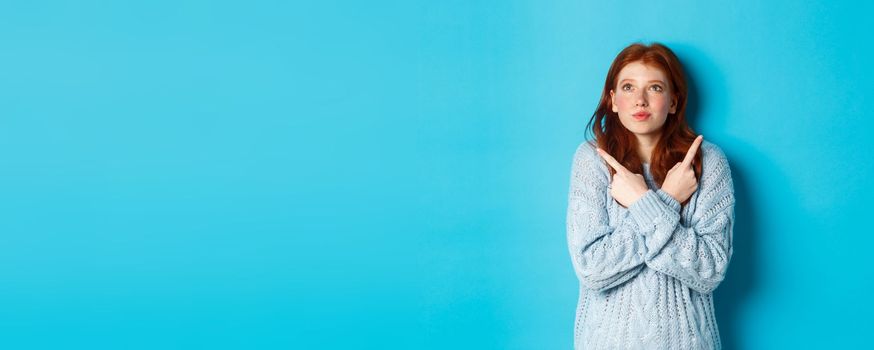 Excited redhead girl pointing fingers sideways, showing two choices and looking tempted up, standing against blue background.