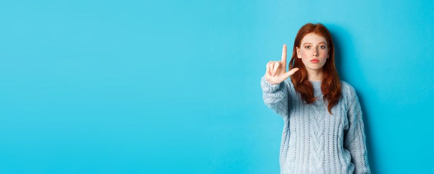 Serious redhead girl in sweater showing taboo gesture, extending one finger, shaking forefinger to disapprove, disagree and forbid something, blue background.