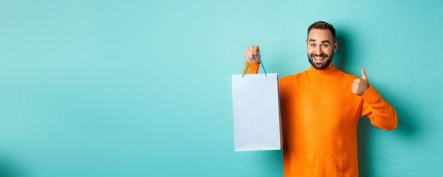 Handsome smiling man showing thumbs-up and shopping bag, recommending store, standing over blue background. Copy space