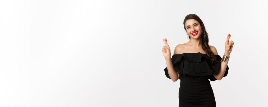 Fashion and beauty. Stylish glamour woman in black dress, red lips, looking optimistic and smiling while cross fingers, making wish, standing over white background.