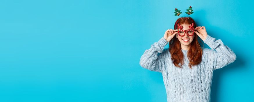 Winter holidays and Christmas sales concept. Beautiful redhead female model celebrating New Year, wearing funny party headband and glasses, smiling at camera.