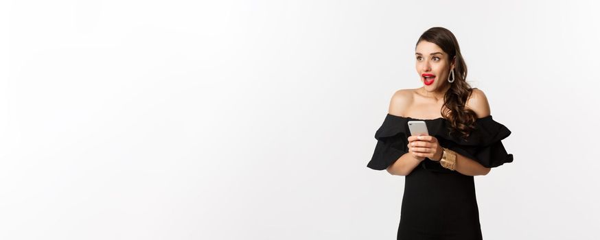Image of excited woman in black glamour dress, red lipstick, using mobile phone and looking left amazed, standing over white background.