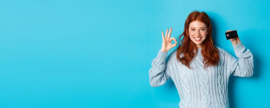 Happy redhead girl in sweater showing credit card and okay sign, recommending bank offer, standing over blue background.