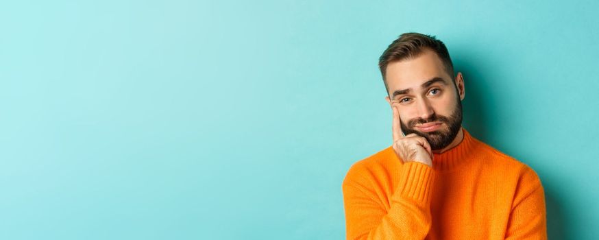 Close-up of unamused sad man looking at camera, pouting and leaning face on hand, standing over light blue background.