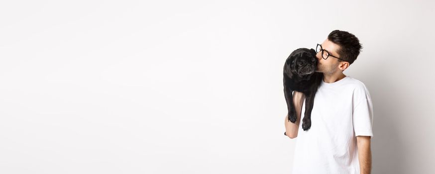 Image of handsome young man kissing his cute black pug, holding dog on shoulder, standing over white background.