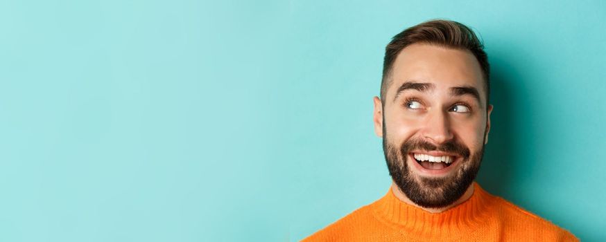 Headshot of handsome caucasian man looking at upper left corner logo and smiling pleased, checking out promo offer, standing over turquoise background.