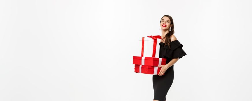 Full length of elegant woman in black dress, red lips, holding christmas presents and smiling pleased, receive gifts, standing over white background.
