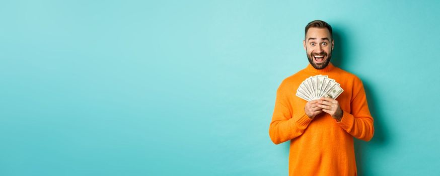 Image of excited bearded guy holding money and rejoicing, winning cash prize, standing against light blue background. Copy space