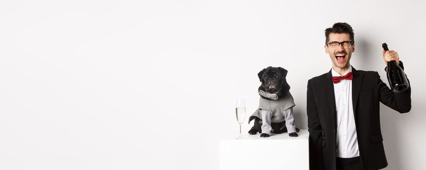 Pets, winter holidays and New Year concept. Happy man celebrating Christmas party pet, standing with cute dog in costume, drinking champagne and rejoicing, white background.