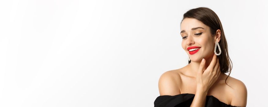 Fashion and beauty concept. Close-up of tender woman in black dress and earrings, gently touching face and smiling, looking down coquettish, standing over white background.