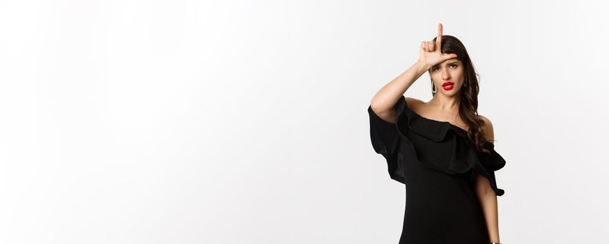 Fashion and beauty. Arrogant glamour woman showing loser sign on forehead, mocking lost person, standing in black dress over white background.