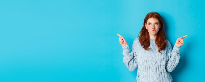 Winter holidays and people concept. Thoughtful redhead girl making decision, pointing fingers sideways, choosing between two ways, standing over blue background.