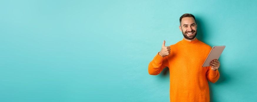 Satisfied adult man smiling, using digital tablet and showing thumb-up, approve and agree, standing against turquoise background.