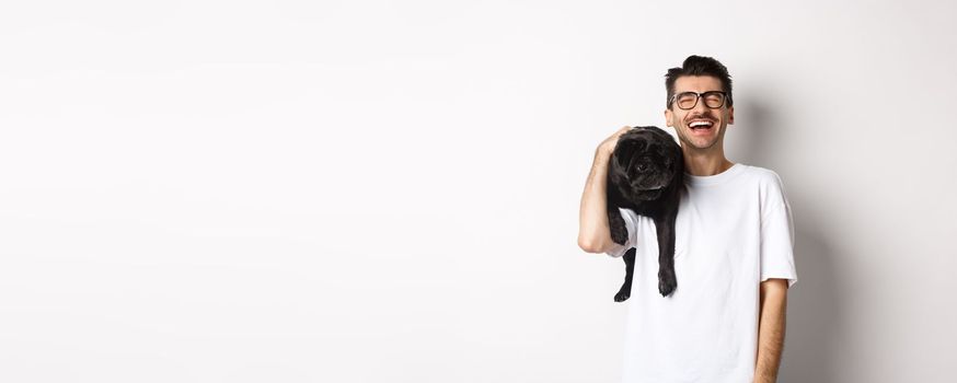 Happy young man laughing carefree, holding cute black dog, pug breed, on shoulder and having fun, standing over white background.