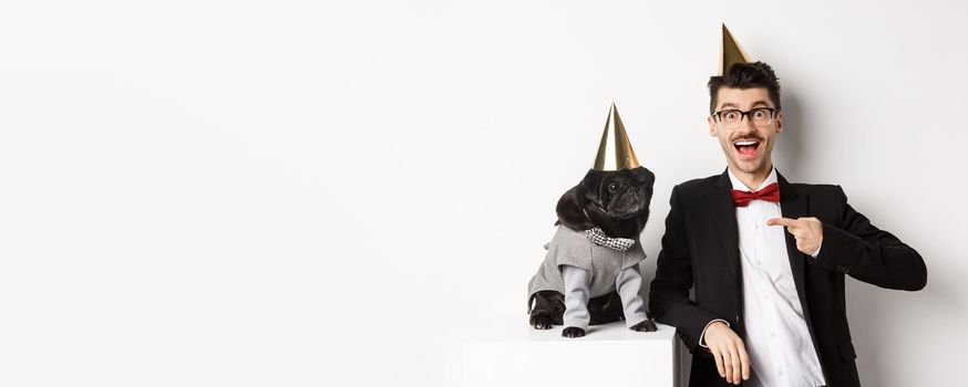 Cheerful man and cute black pug wearing party cones and suits, dog owner celebrating pet birthday, standing over white background.