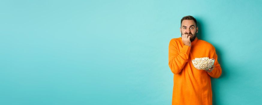 Image of scared young man watching horror movie, biting fist and looking terrified, holding bowl of popcorn, standing over turquoise background.