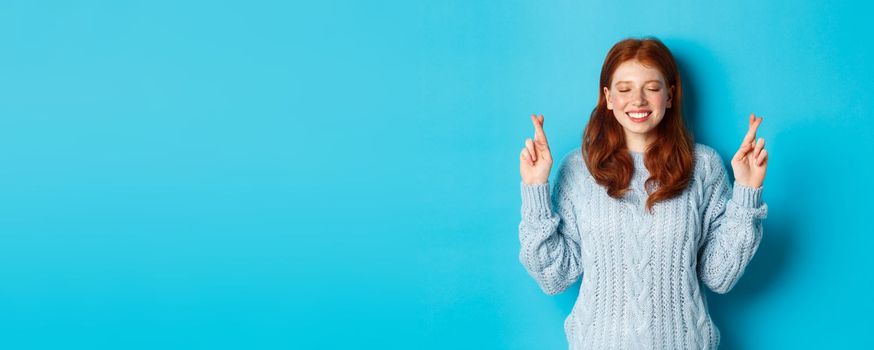 Hopeful redhead girl making a wish, cross fingers for good luck, smiling and anticipating good news or positive result, standing against blue background.