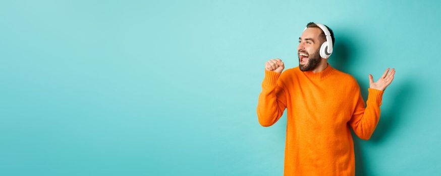 Happy man lipsync while listening music in headphones, holding invisible microphone, standing over turquoise background.