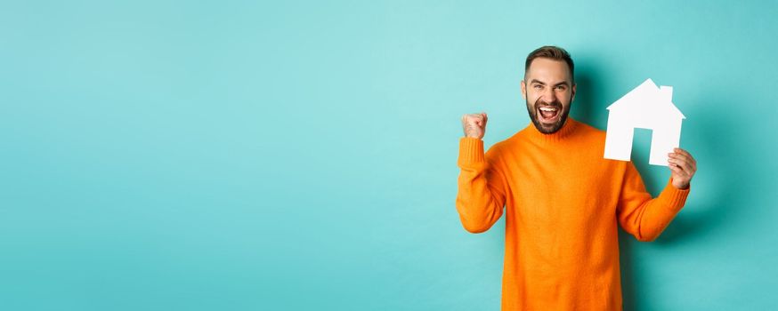Real estate concept. Excited man saying yes, showing paper house maket and looking satisfied, standing in orange sweater over light blue background.