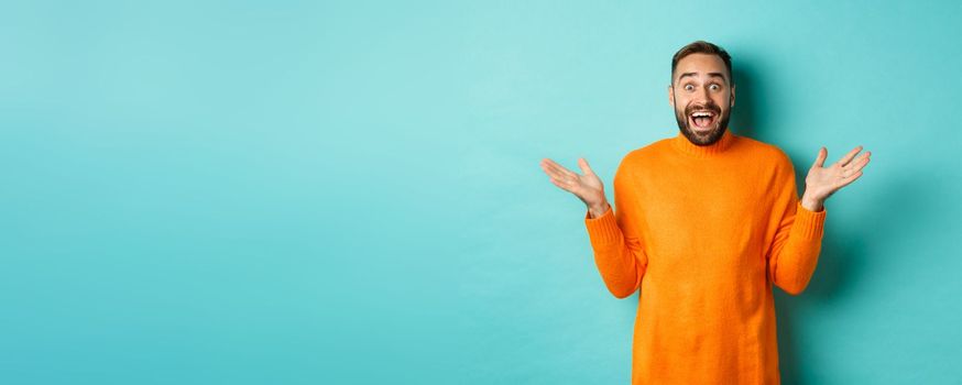 Surprised man raising hands up, looking at something amazing, standing over turquoise background in winter sweater.