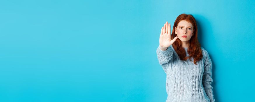 Serious and confident redhead girl telling to stop, saying no, showing extended palm to prohibit action, standing over blue background.