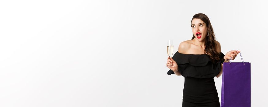 Excited woman shop and drink champagne, holding shopping bag, looking amazed, standing against white background.