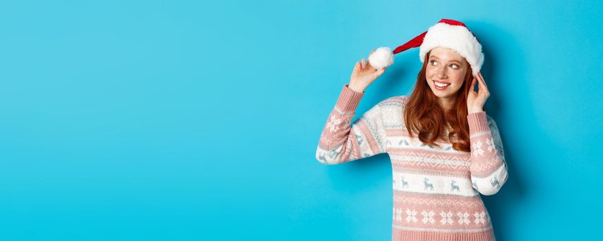 Winter and Christmas Eve concept. Cute teenage girl wearing santa hat and sweater, celebrating xmas, smiling happy, standing over blue background.