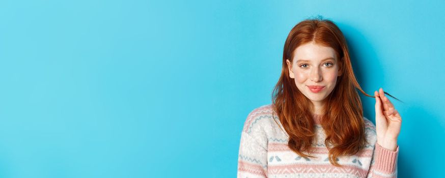 Close-up of confident and sassy redhead teen girl looking at camera pleased, playing with hair strand and smirking, standing over blue background.