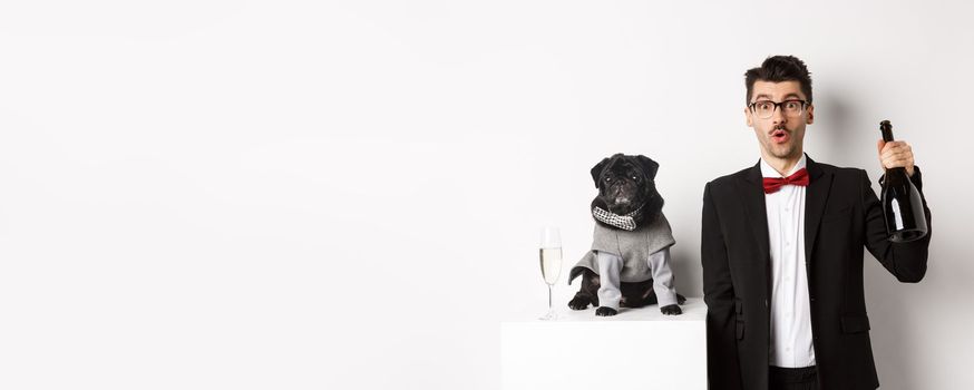 Pets, winter holidays and New Year concept. Cheerful man with cute black pug dog celebrating Christmas party, holding champagne bottle and smiling, white background.