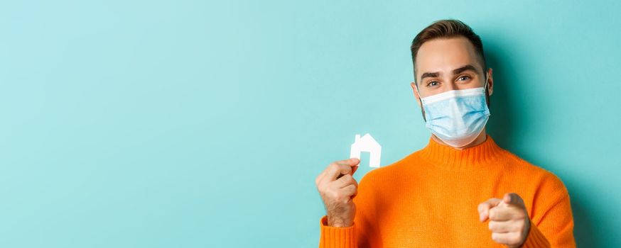 Real estate and coronavirus pandemic concept. Close-up of adult man in medical mask pointing at you and showing small paper house, standing over light blue background.