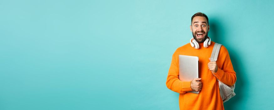 Happy man with backpack and headphones, holding laptop and smiling, looking excited, standing over turquoise background.