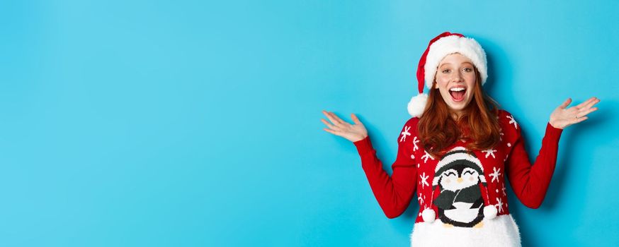 Happy holidays concept. Cheerful redhead girl in xmas sweater and santa hat, raising hands up and wishing merry Christmas, standing against blue background.