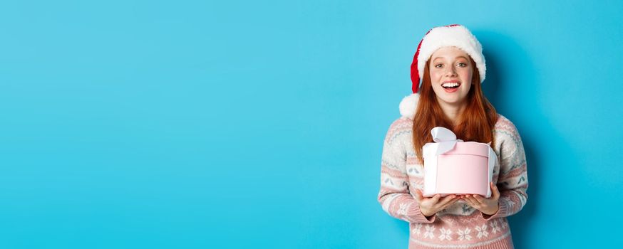 Winter and celebration concept. Happy redhead girl receiving christmas gift and thanking, hugging box with presents and smiling, standing in santa hat over blue background.