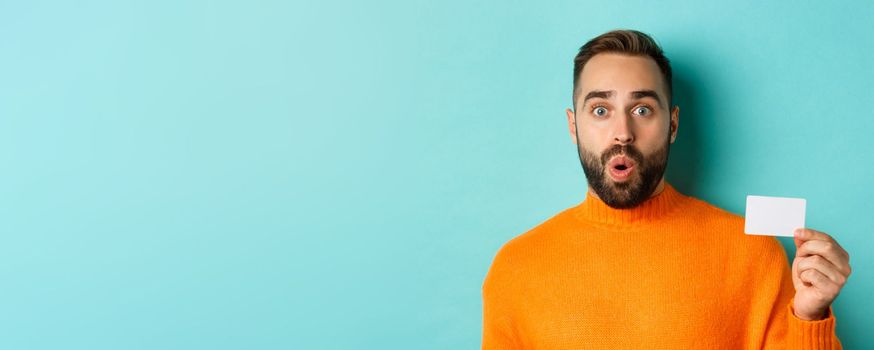 Close-up of handsome caucasian man going on shopping, showing credit card and smiling, standing over turquoise background.