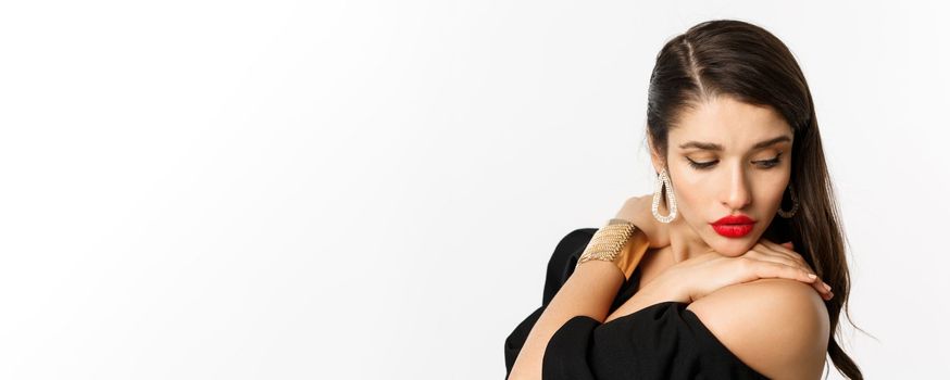 Fashion and beauty concept. Close-up of sensual woman in elegant earrings and black dress, wearing makeup with red lips, looking down tenderly, standing over white background.