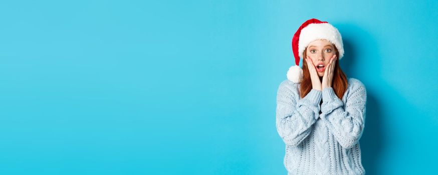 Winter holidays and Christmas Eve concept. Surprised redhead girl in santa hat, staring with disbelief at camera, open mouth amazed, standing over blue background.