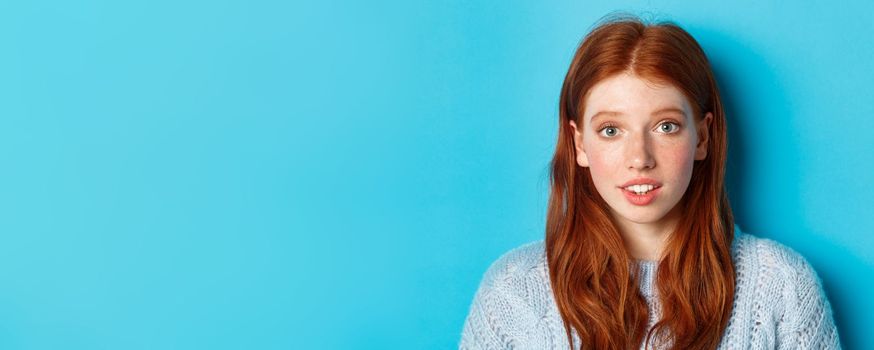 Headshot of cute redhead girl with freckles, looking hopeful and innocent at camera, standing over blue background.