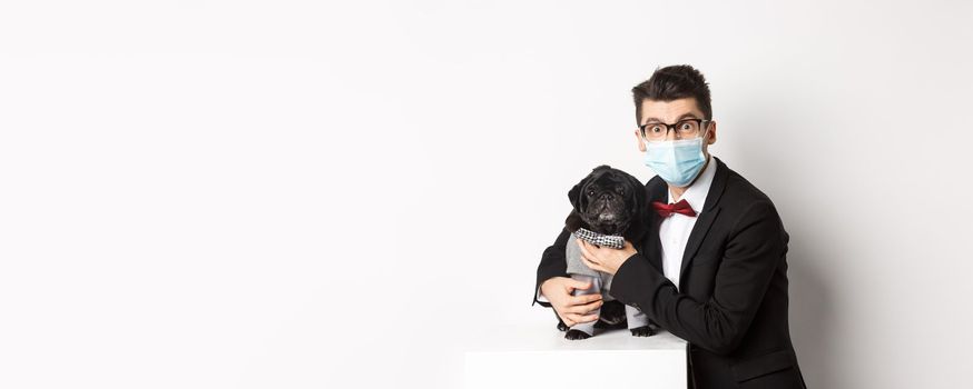 Coronavirus, pets and celebration concept. Happy dog owner in suit and face mask hugging cute black pug in costume, standing over white background.