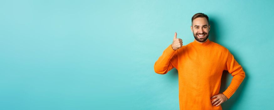 Well done. Handsome bearded man showing thumb up, praising good work, recommend excellent product, like and approve, smiling satisfied, standing over light blue background.