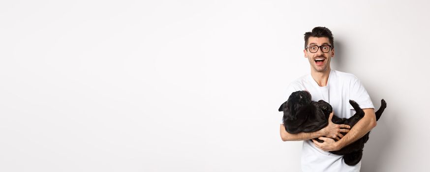 Handsome young man playing with cute black puppy. Dog owner petting a pug, standing over white background.